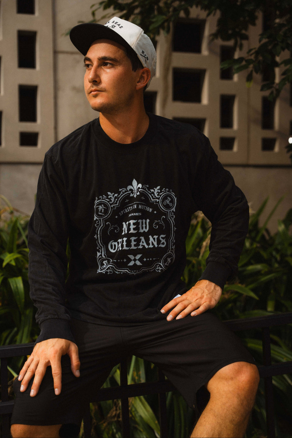 A man wearing a black long-sleeve t-shirt with a white 'New Orleans' graphic print on the front, paired with black shorts and a white baseball cap, sitting outdoors in front of a building wall and green plants.